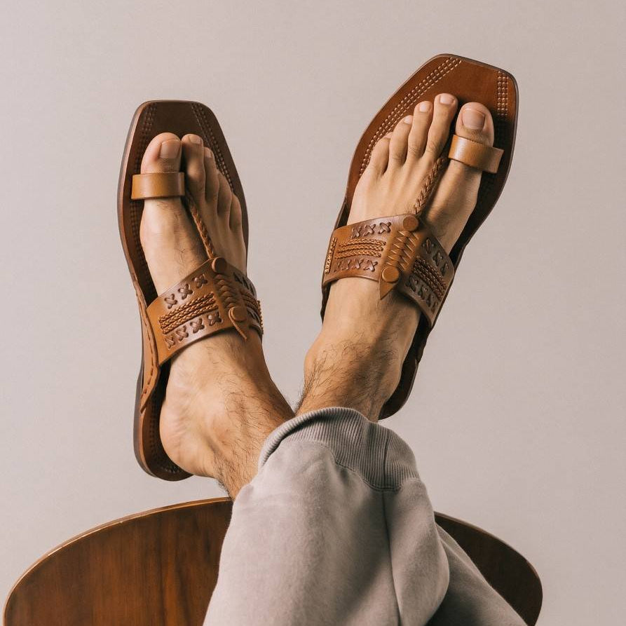 A person wearing brown leather Kolhapuri chappal with woven detailing, shown from above while sitting on a wooden chair against a plain light gray background.